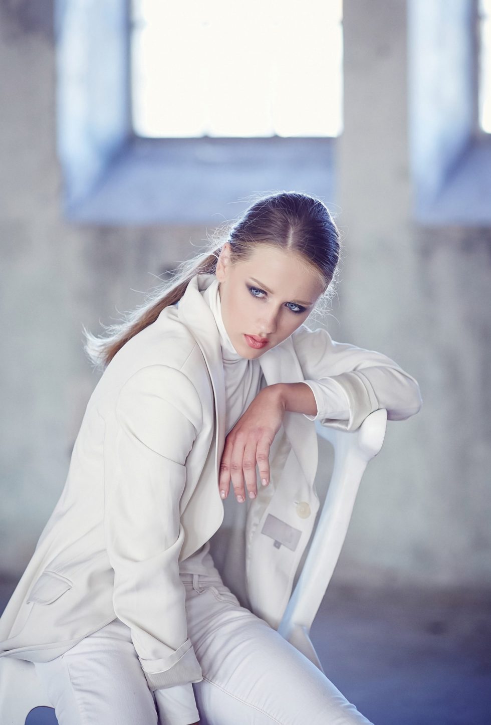 Blue eyes girl in a white clothes sits on a chair in the room with big window.